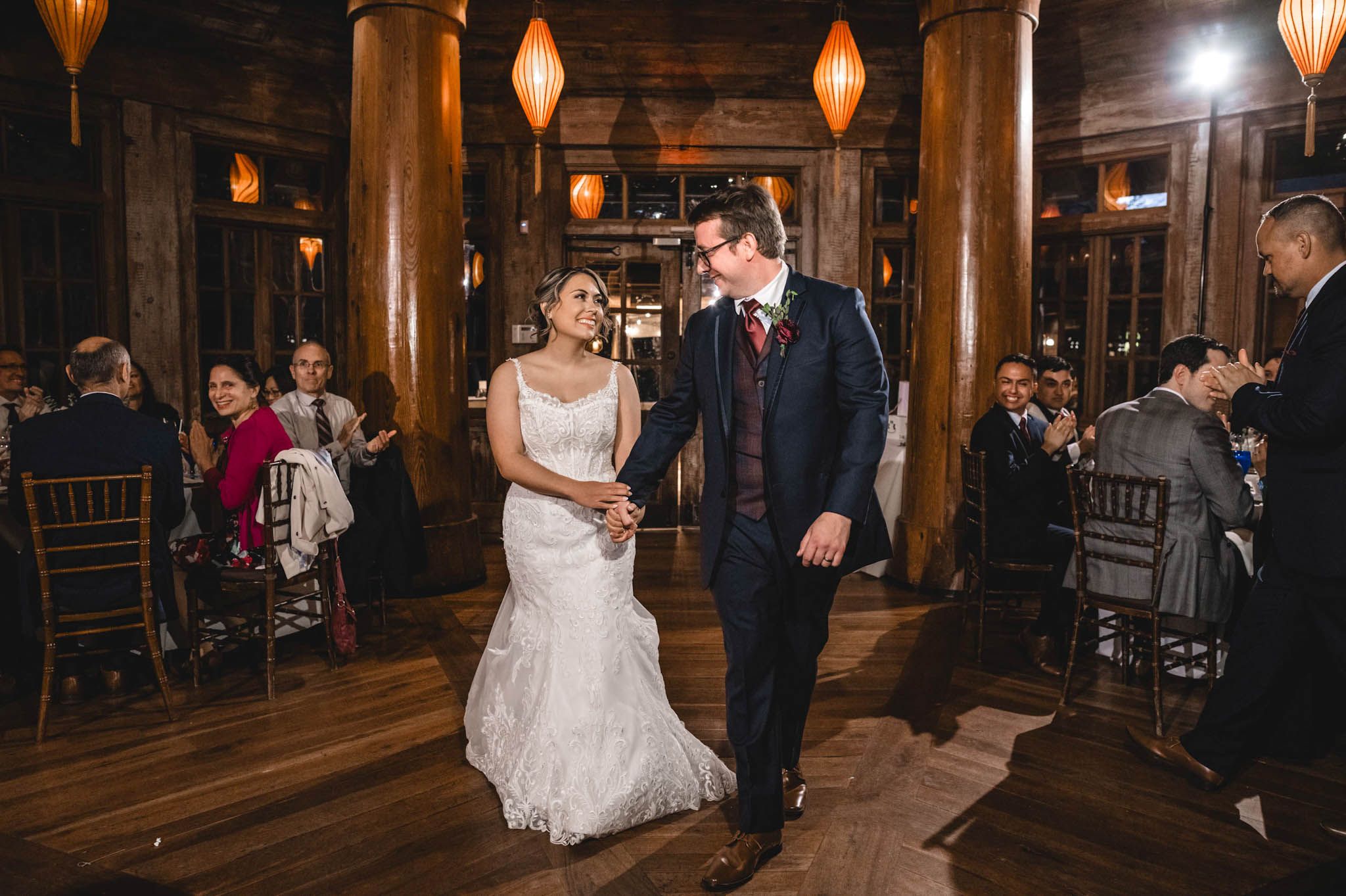 Wedding reception guests dancing in a rustic wooden venue with warm glowing pendant lights overhead.