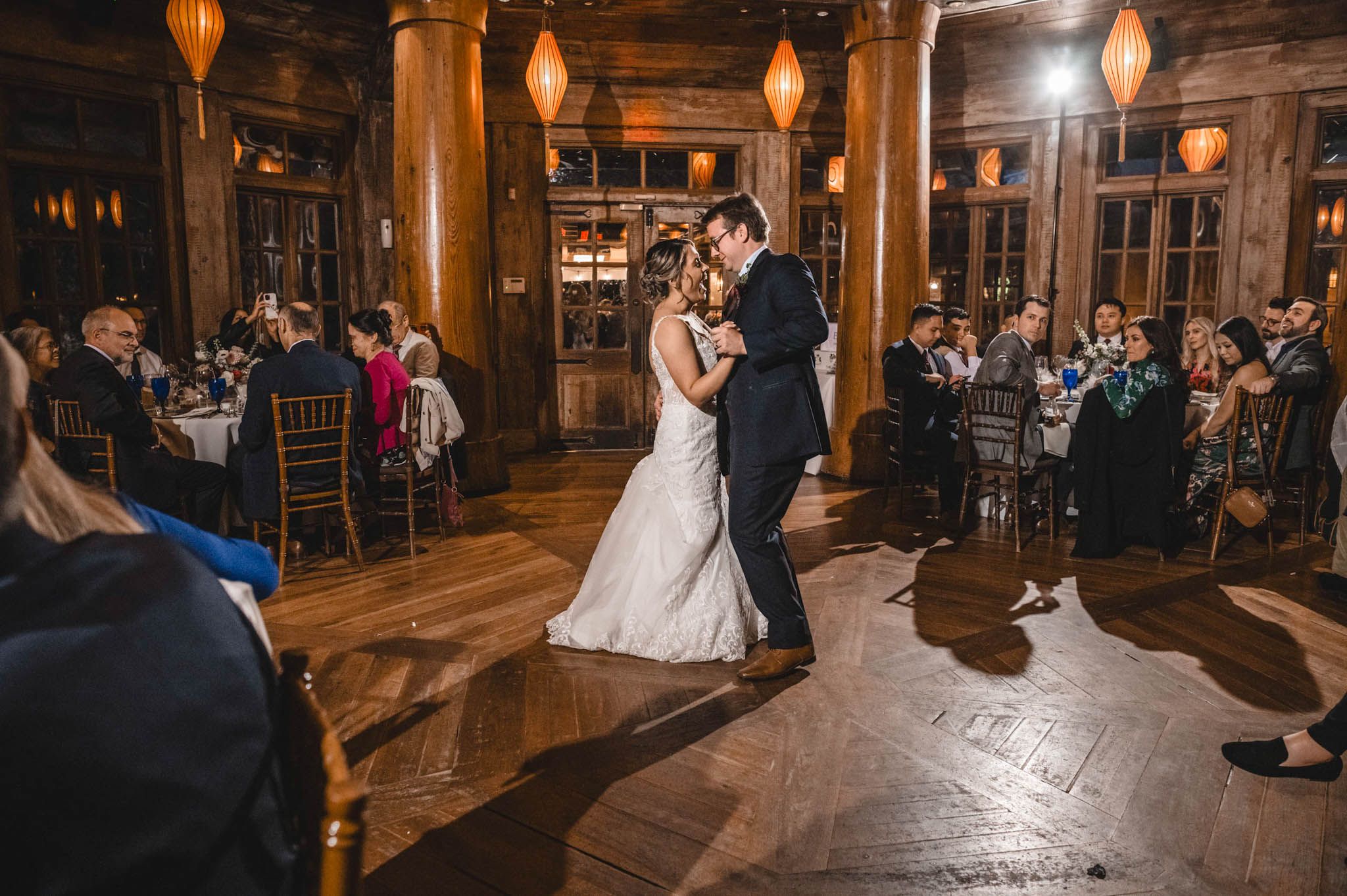 Wedding party dancing on wooden floors under vintage pendant lights in rustic reception hall.