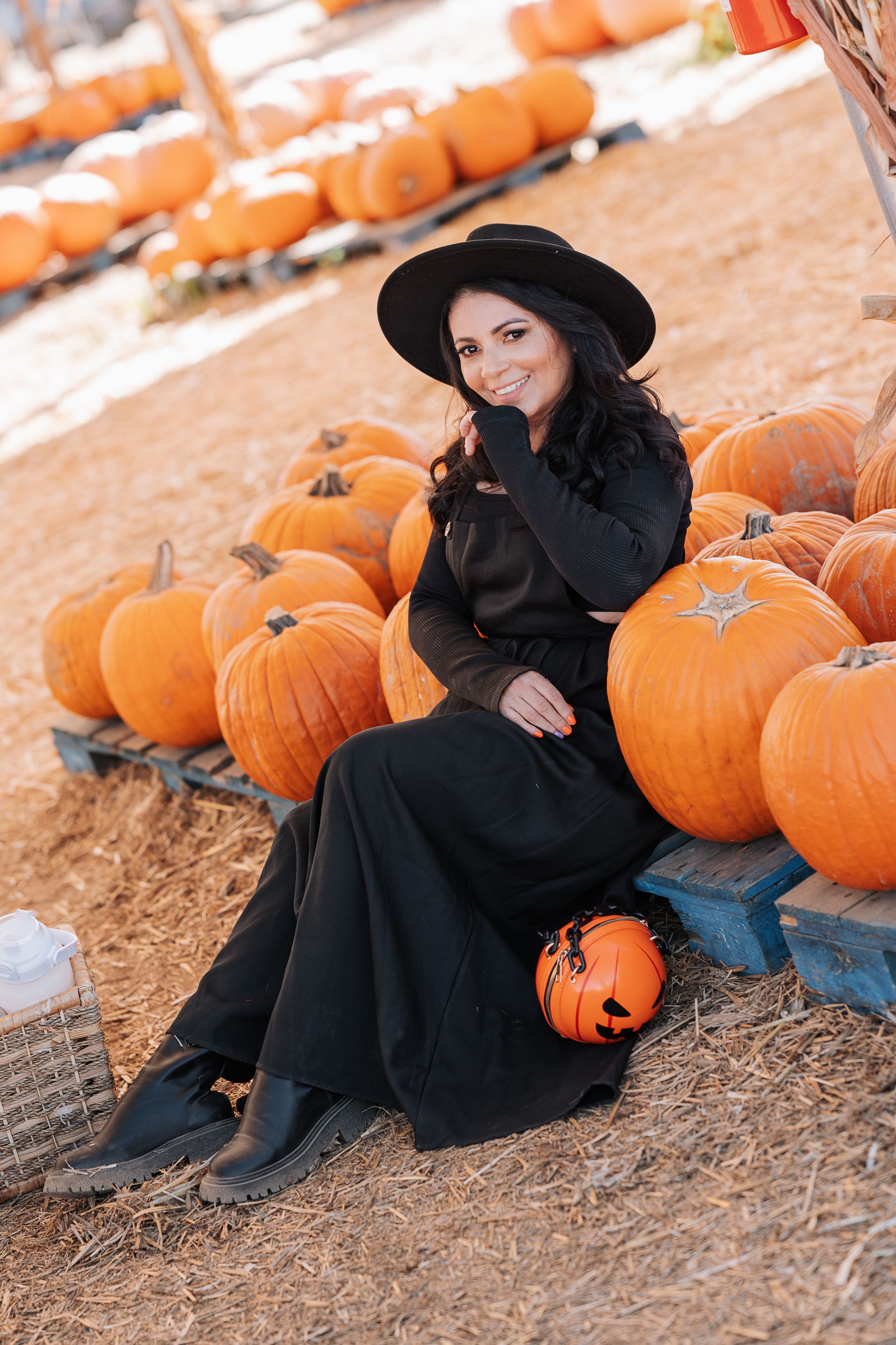 A person in dark attire sits among pumpkins at a seasonal pumpkin patch.