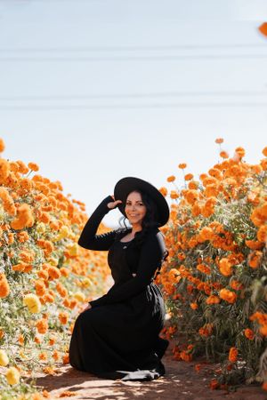 A figure in a black dress and wide-brimmed hat poses among vibrant orange flowers in a garden setting.