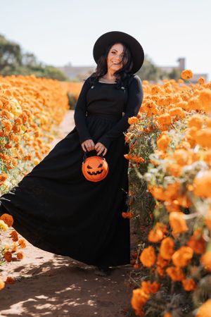 A figure in gothic attire holds an orange pumpkin basket while standing among orange marigolds.