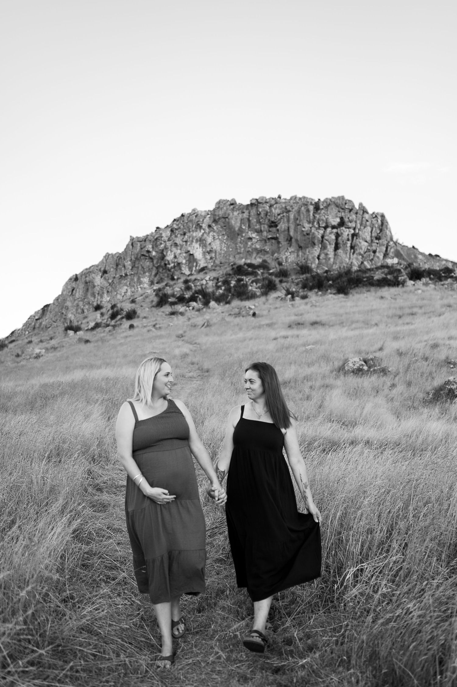 Two women in flowing dresses walk together through tall grass with a rocky outcrop in the background.
