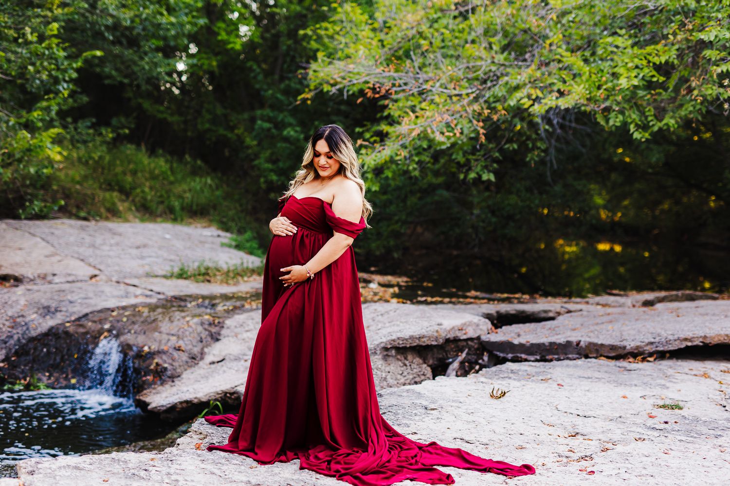 Maternity photo of person in flowing burgundy dress standing by waterfall in nature setting.