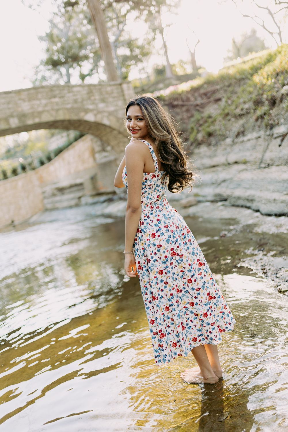A woman in a floral dress splashes and plays in a shallow stream on a sunny afternoon.