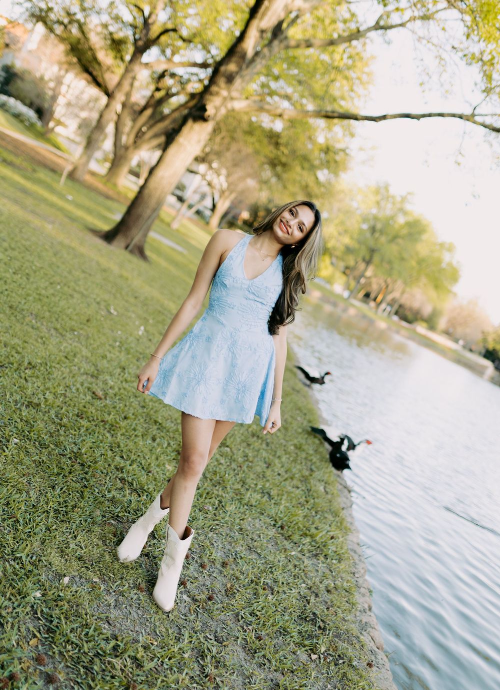 Someone wearing a light blue dress stands in a park setting with autumn trees and a lake in the background.