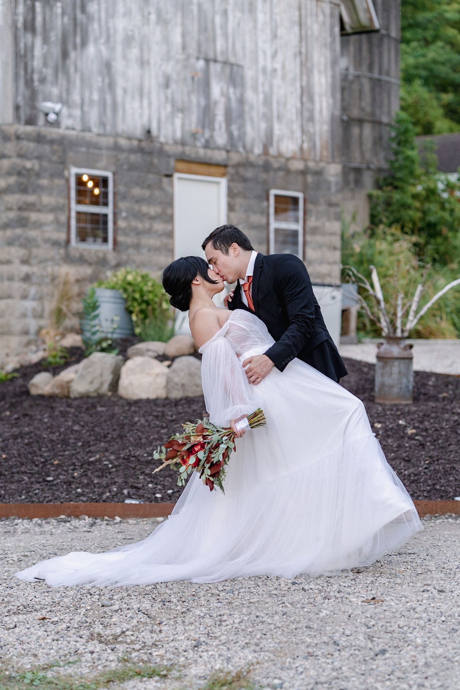 A groom dips his bride for a romantic kiss outside a rustic barn, as she holds a red floral bouquet in her white gown.