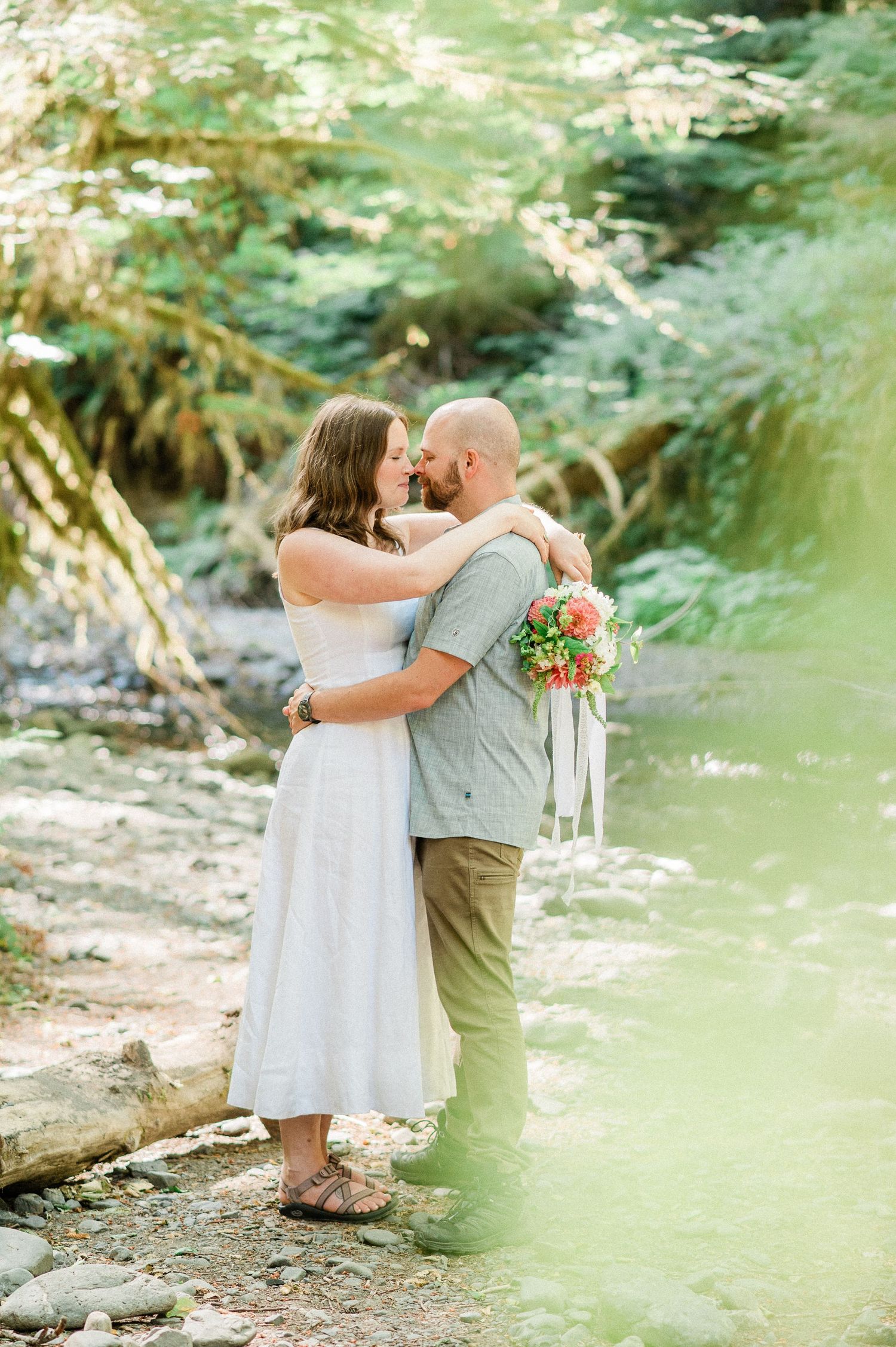 A romantic outdoor photo of a couple kissing by a stream in a lush forest setting with a colorful bouquet.