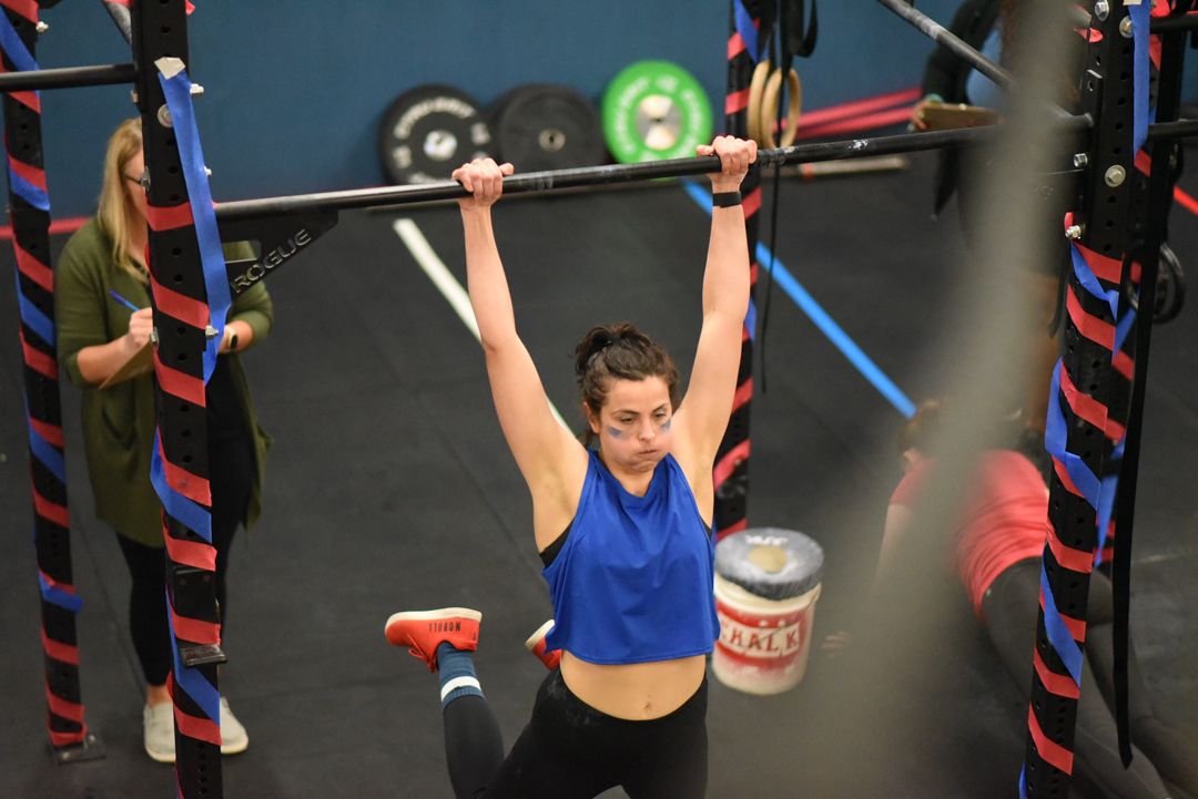 Athlete in blue tank top performs an overhead lift at a CrossFit gym with fitness equipment visible in background.