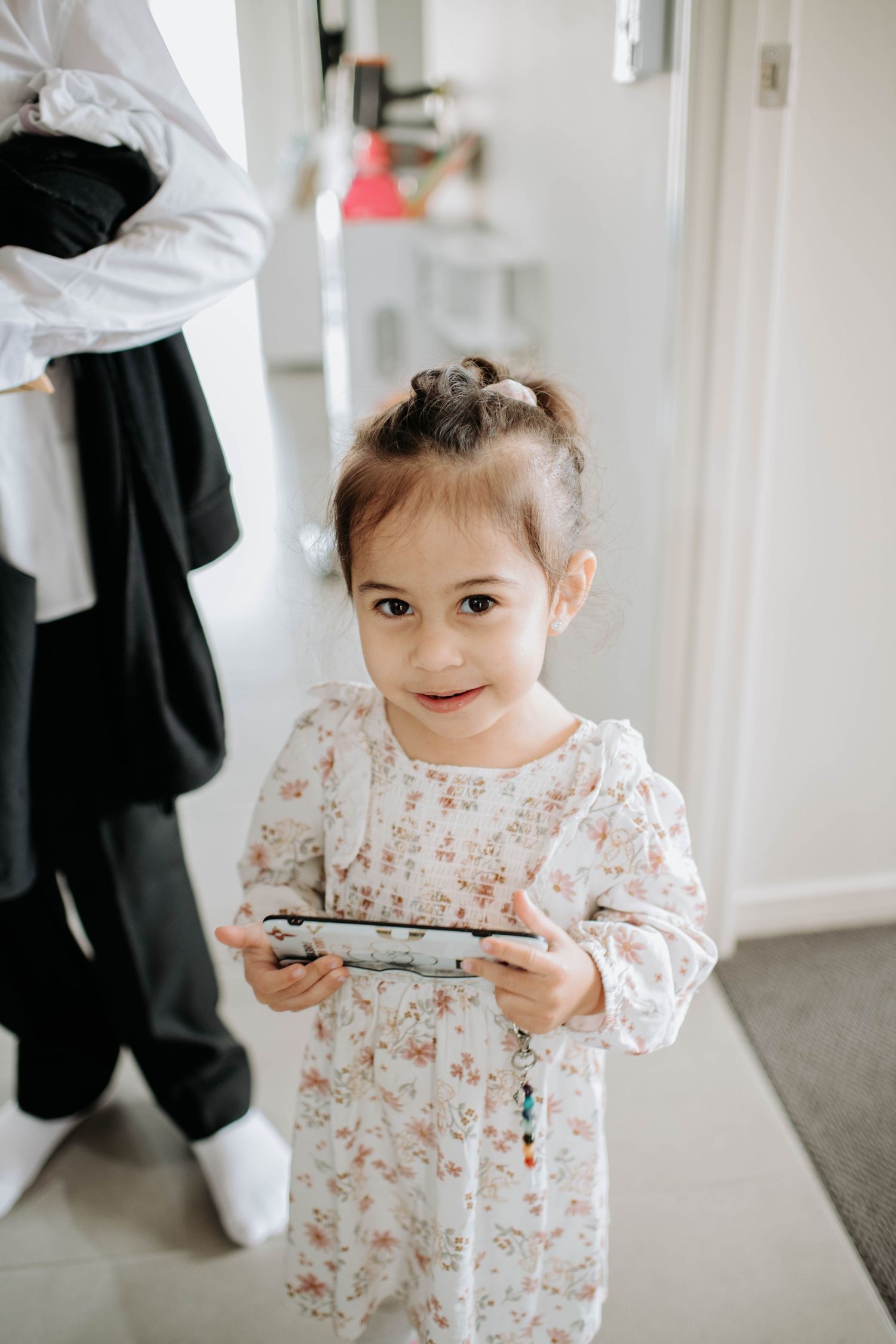 A small child in a floral dress holds a phone during wedding preparations.