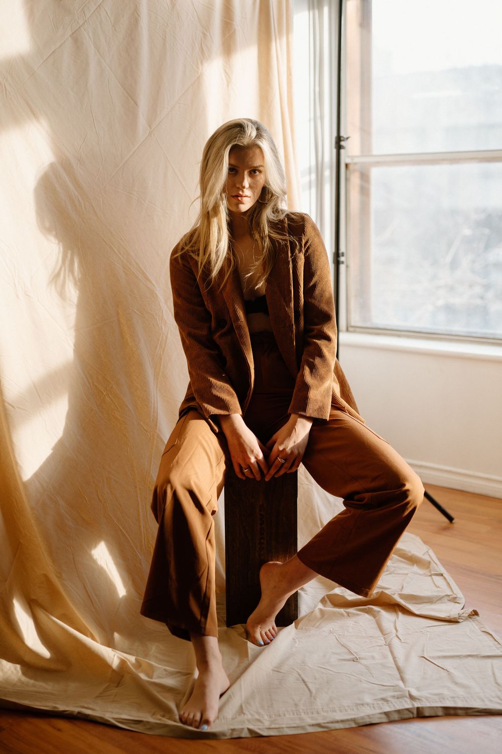 Person in brown attire sitting on wooden box by window with natural sunlight streaming in.