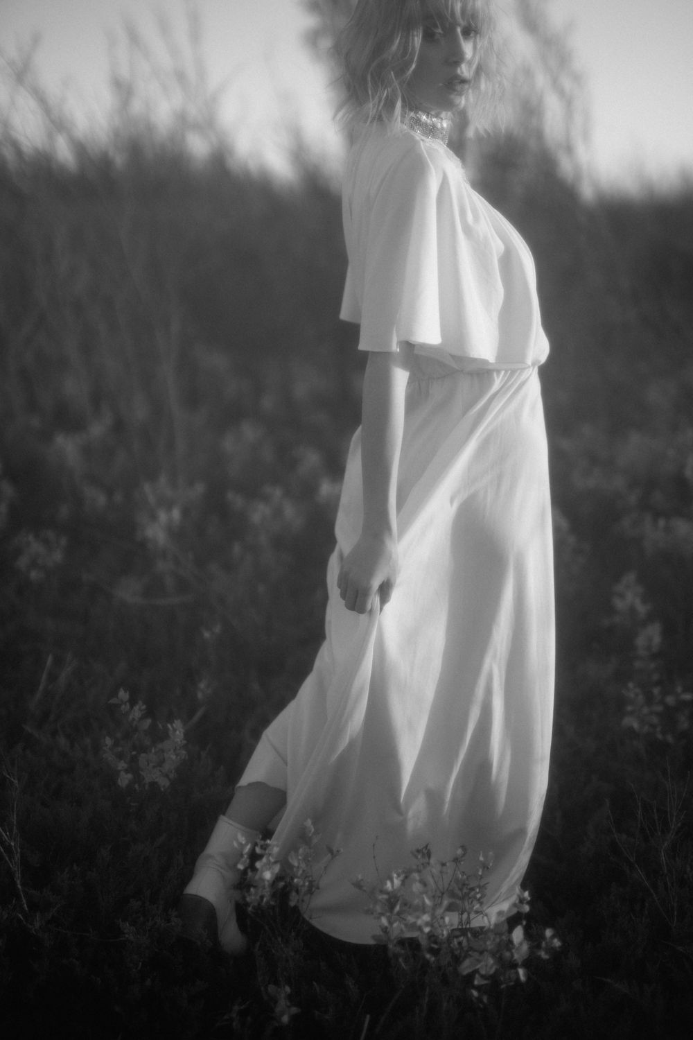 Dreamy black and white photo of figure in flowing white dress standing among wildflowers.