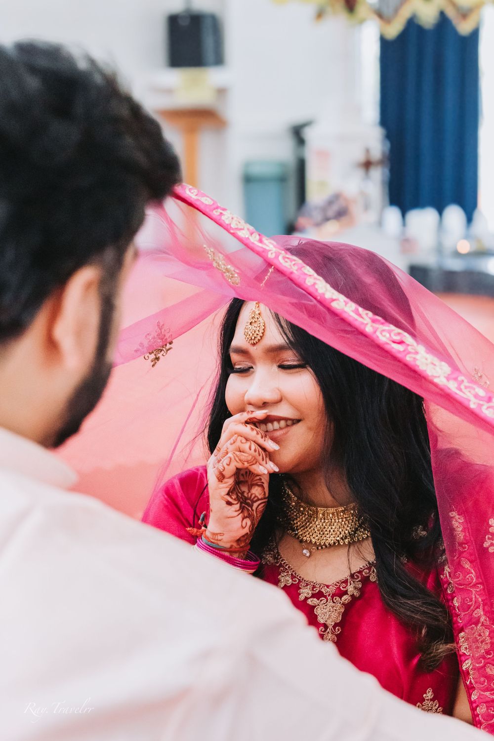 Bride in traditional red attire smiles under pink veil during wedding ceremony.