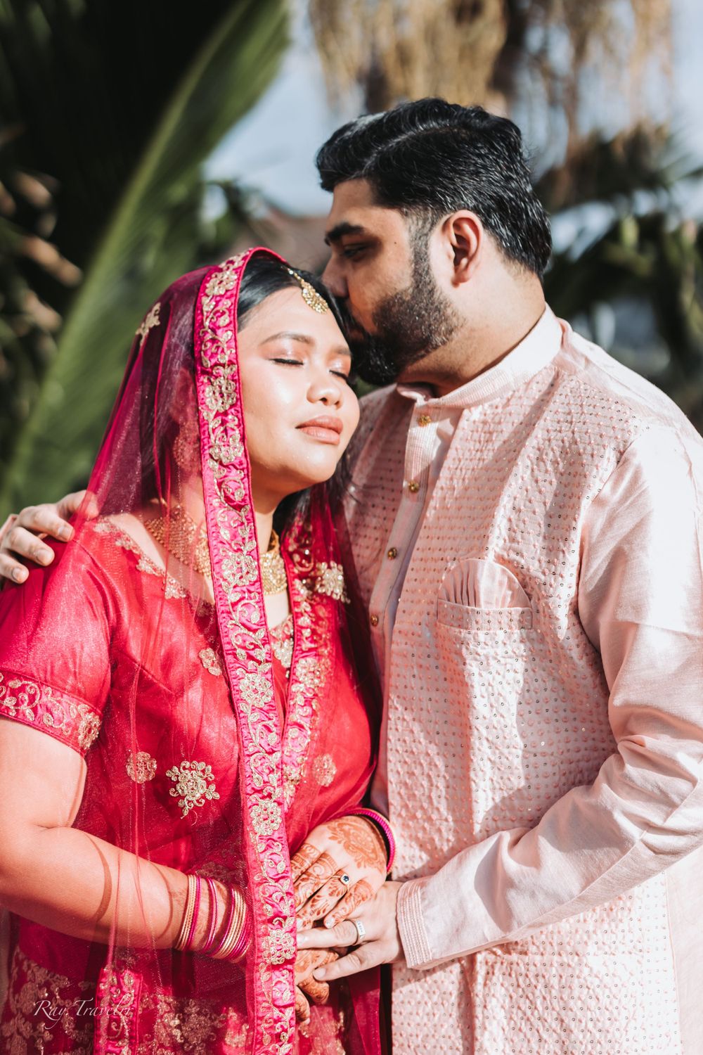 Intimate moment between couple in traditional South Asian wedding attire under palm trees.