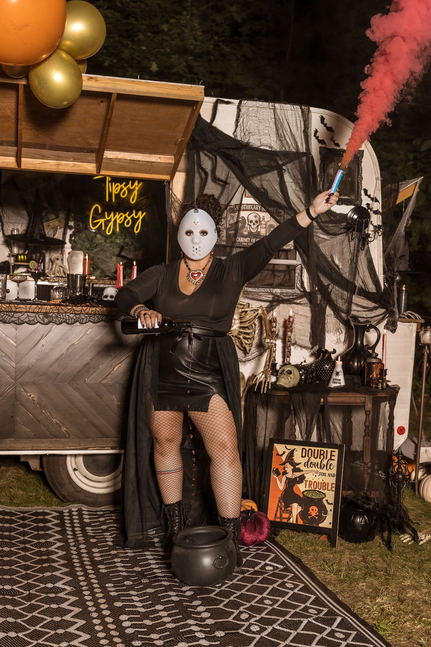 A figure in Halloween costume with white mask poses next to orange and gold balloons at outdoor party setup.