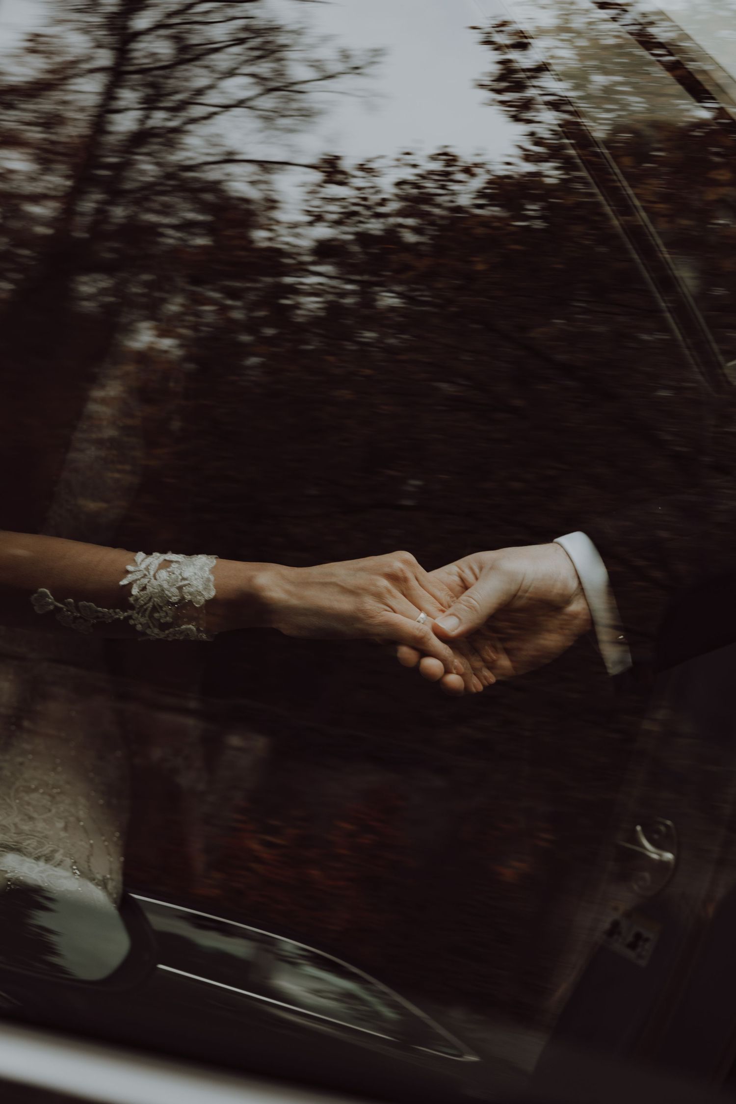 Close-up of newlywed couple holding hands through car window with trees in background.