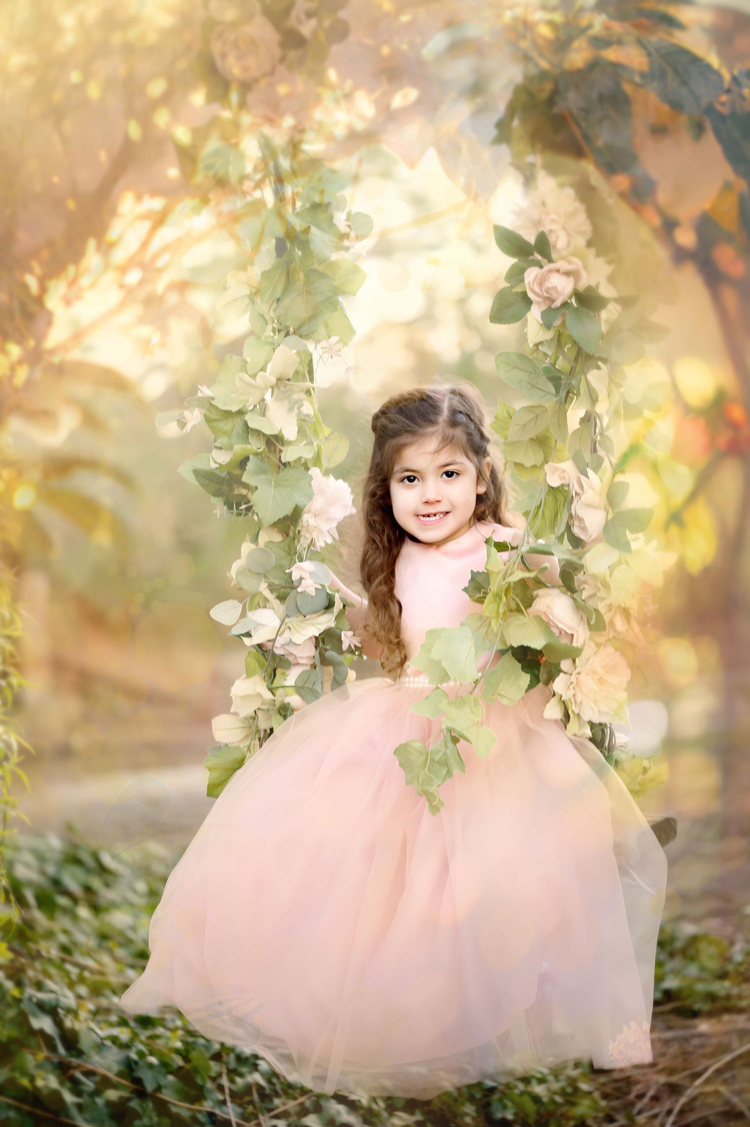 Child in pink princess dress sits on floral swing in magical garden setting.