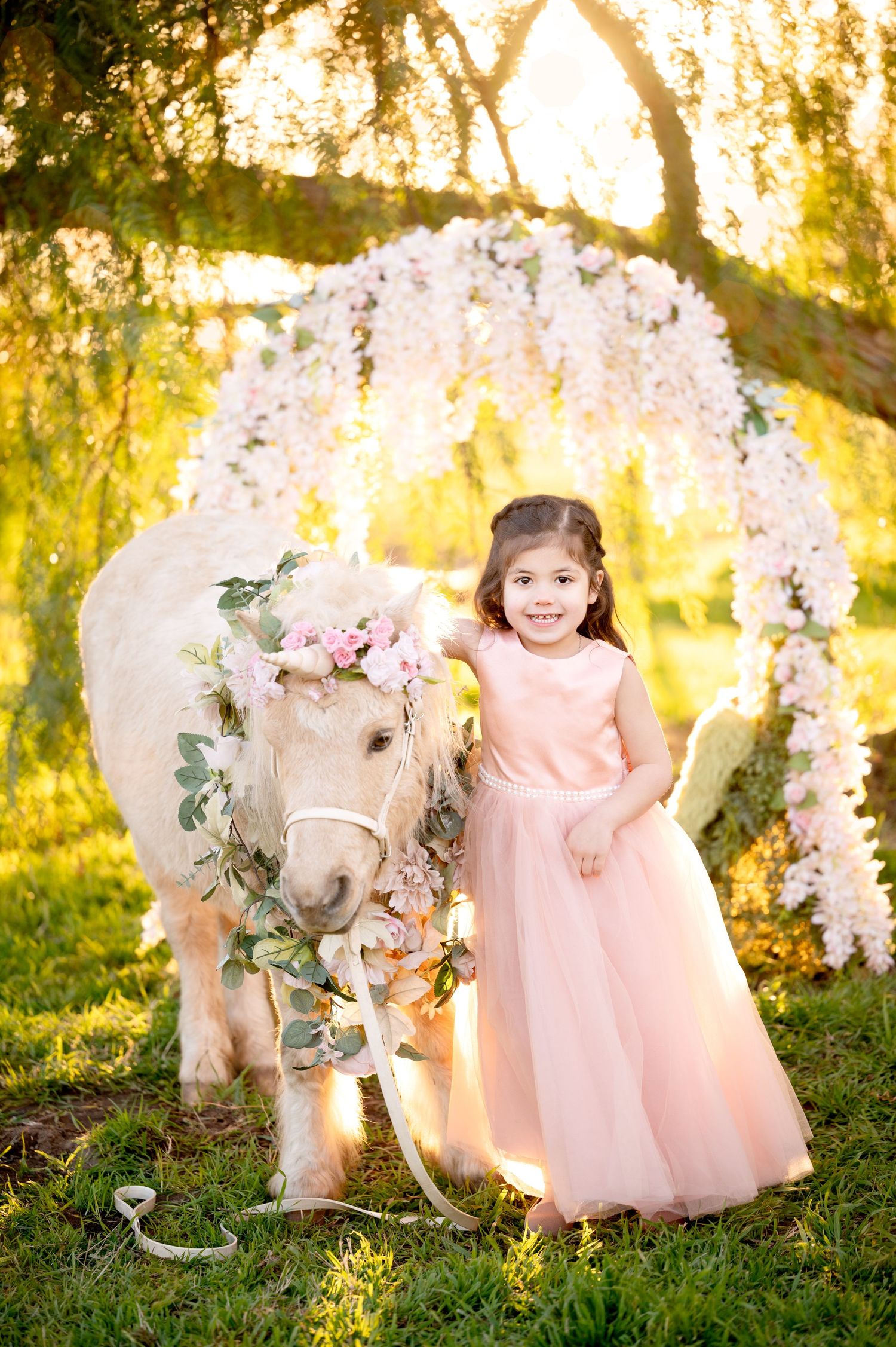 Sunlit garden scene with decorated white pony and pink tulle dress under floral arch.