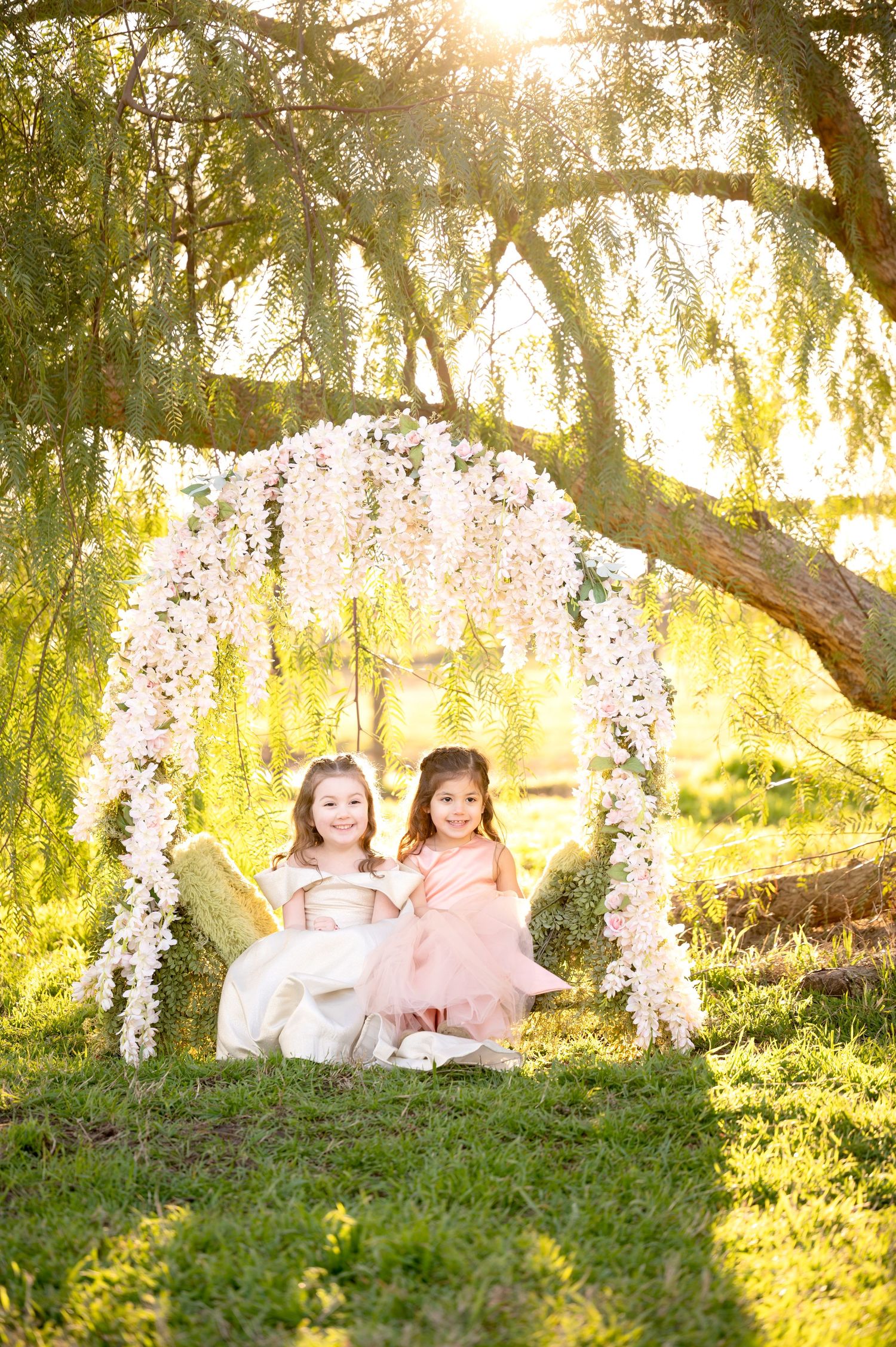 Two children in pink and white dresses sit under a floral arch as golden sunlight filters through tree branches.