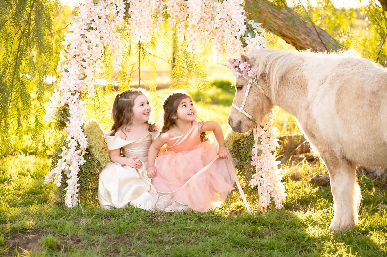 Children and white pony share magical moment under hanging wisteria in golden sunset light.