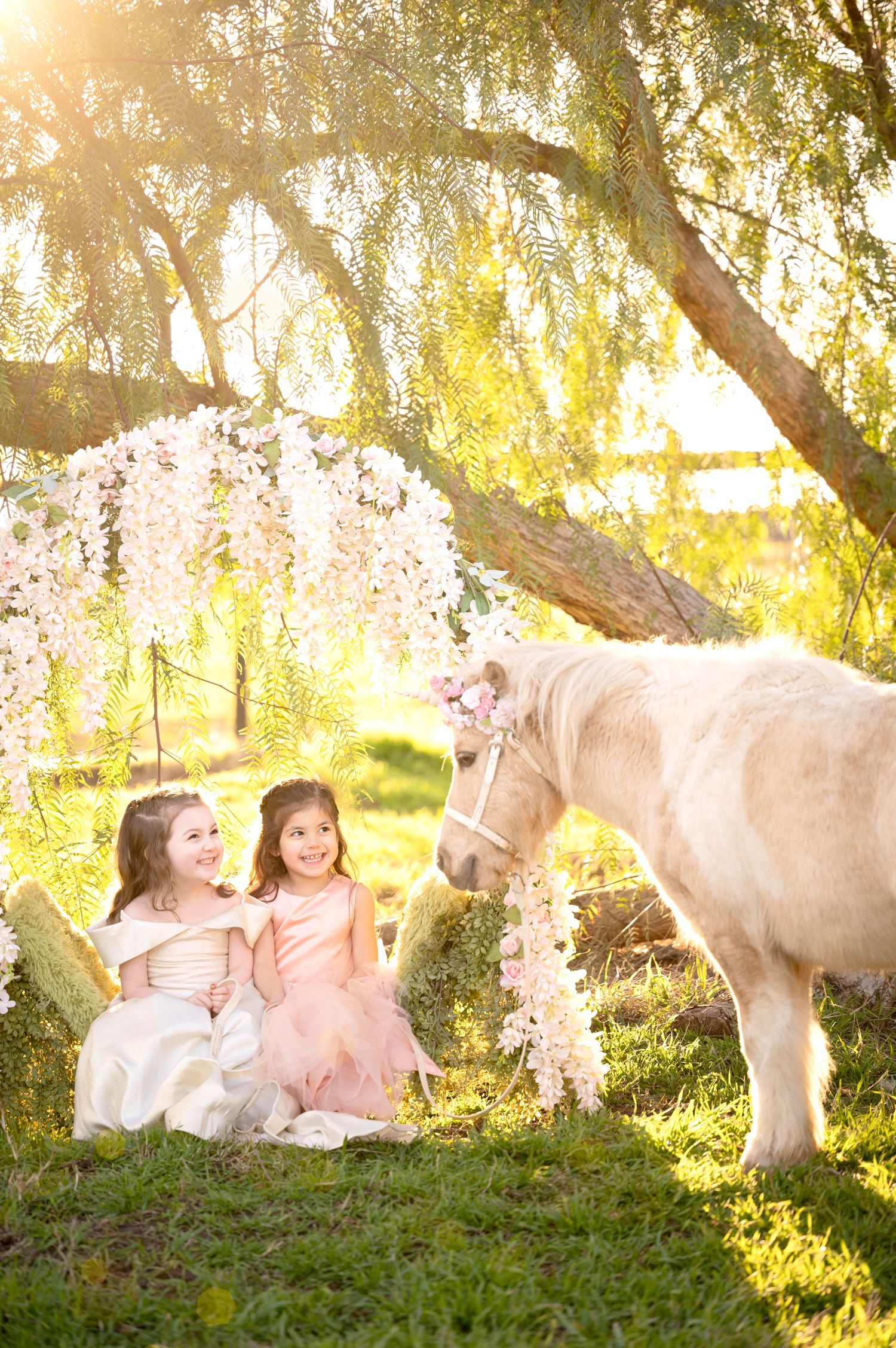 A magical scene with a white pony visiting two children in fancy dresses under a flower-decorated willow tree.