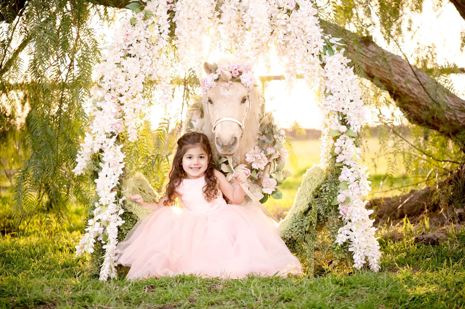 Whimsical photo of pink tulle dress under floral arch with decorated pony in dreamy garden setting.