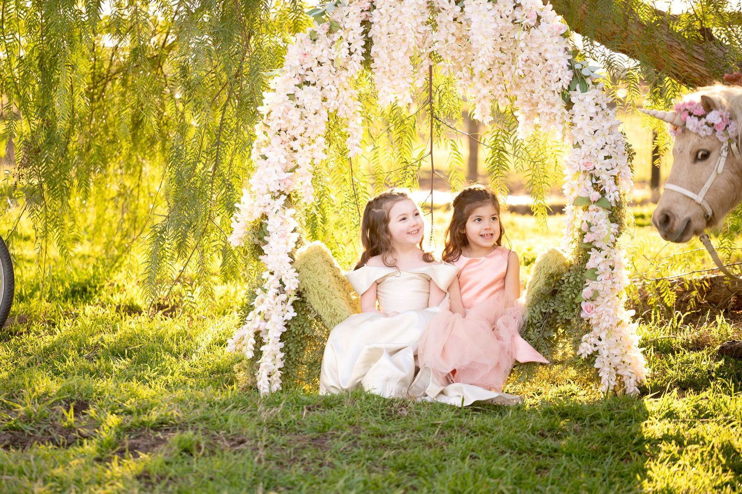 Two children in tulle dresses sit on a floral swing beneath a willow tree with a white pony beside them.