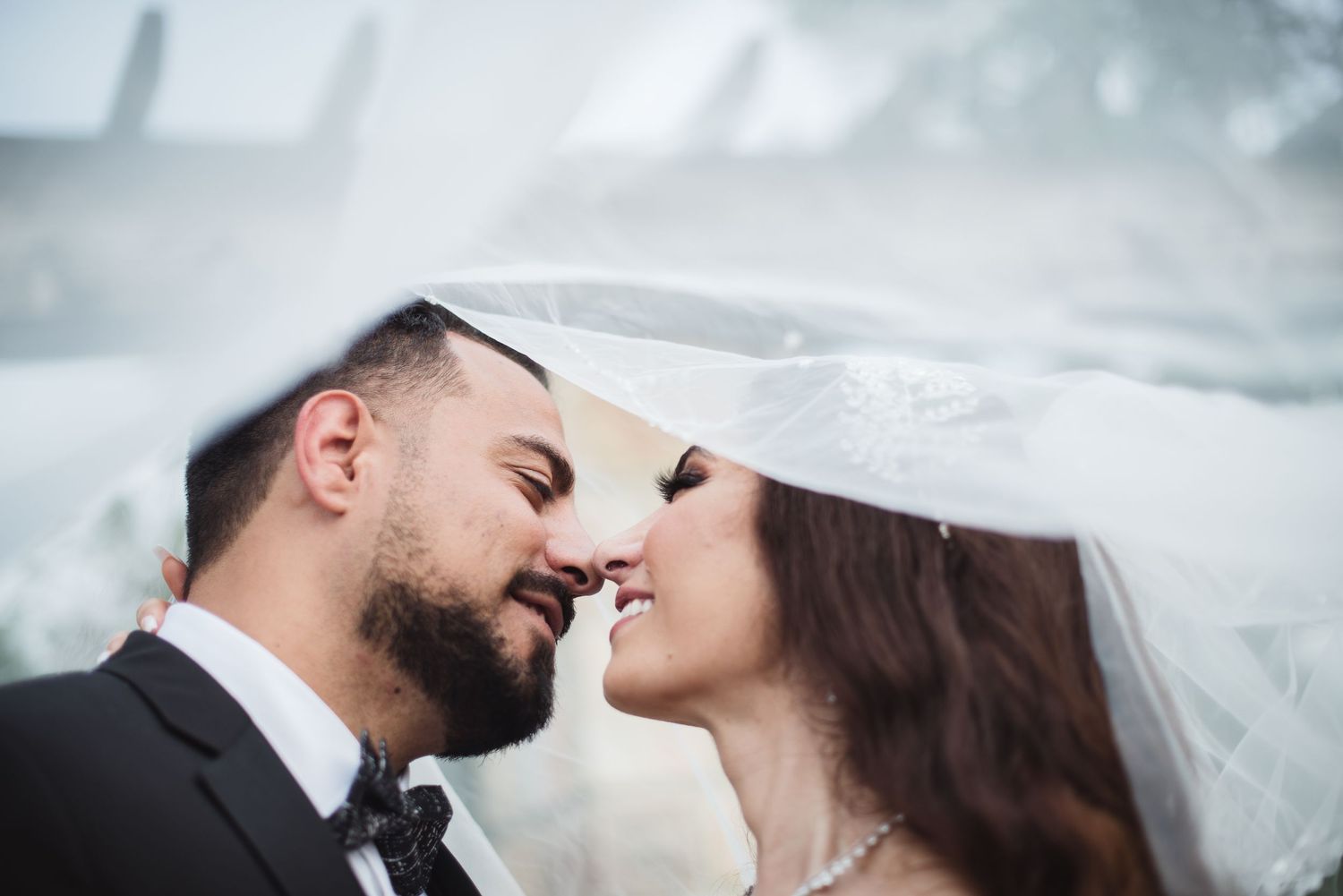 Bride and groom share a romantic nose-to-nose moment under a flowing white wedding veil.