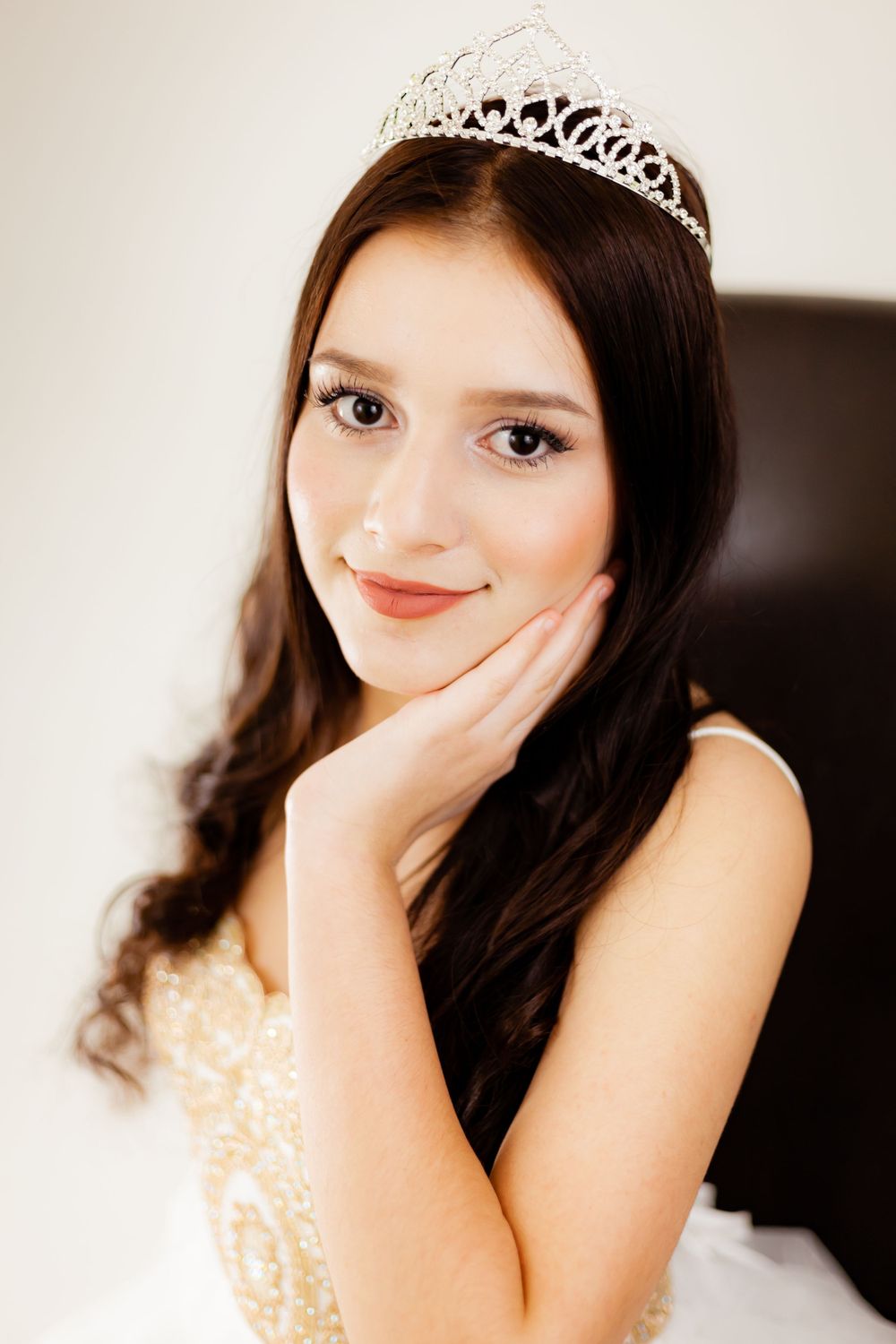 Graceful pose in formal attire with crystal tiara against neutral backdrop.