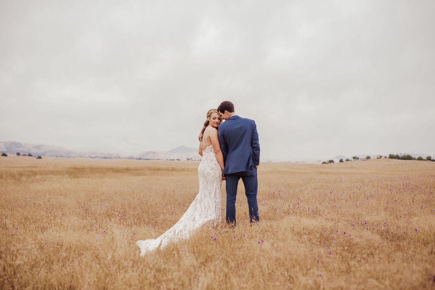 Newlyweds standing together in golden wheat field looking at distant horizon.
