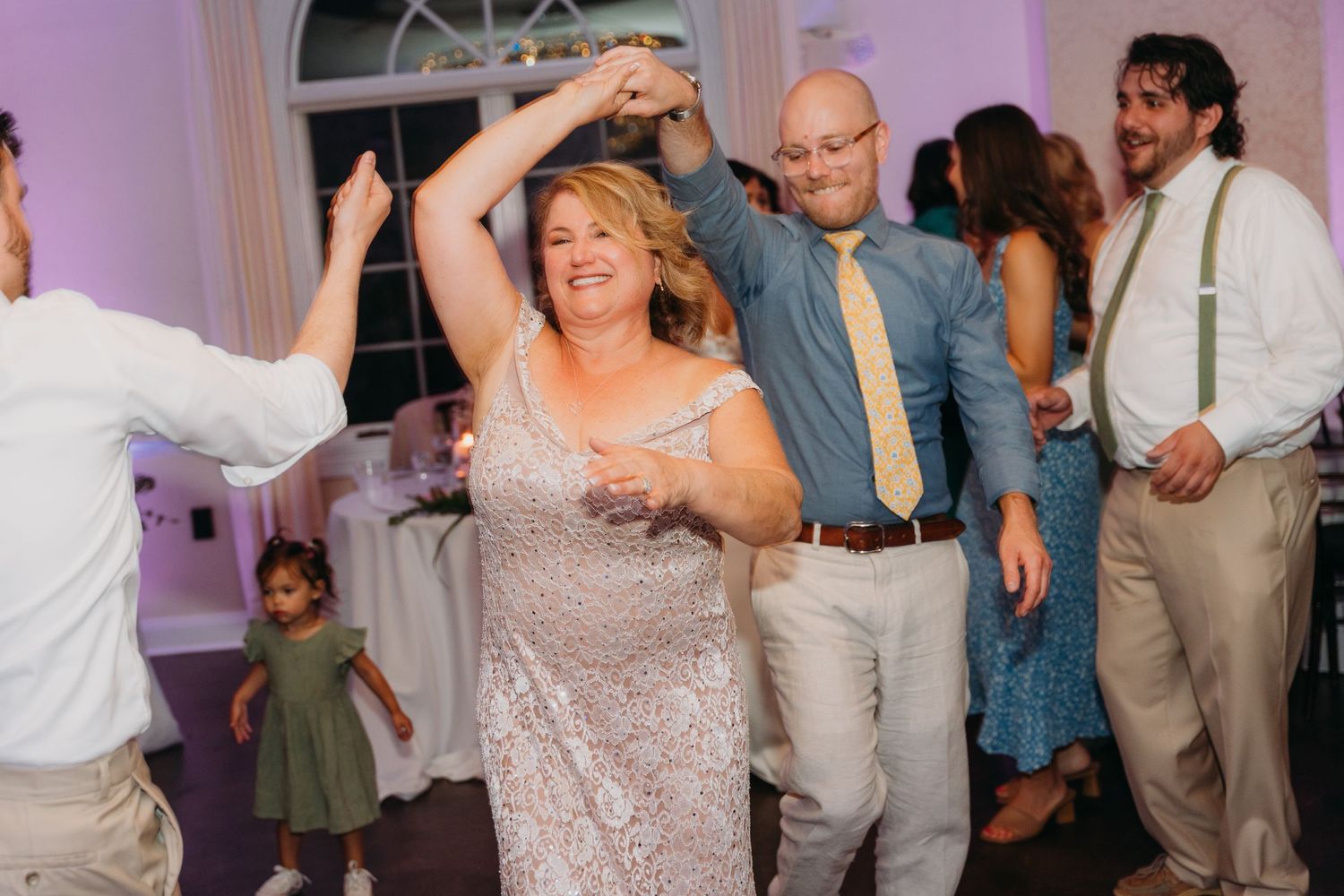 Wedding guests dancing and celebrating on the dance floor at a reception with purple lighting.