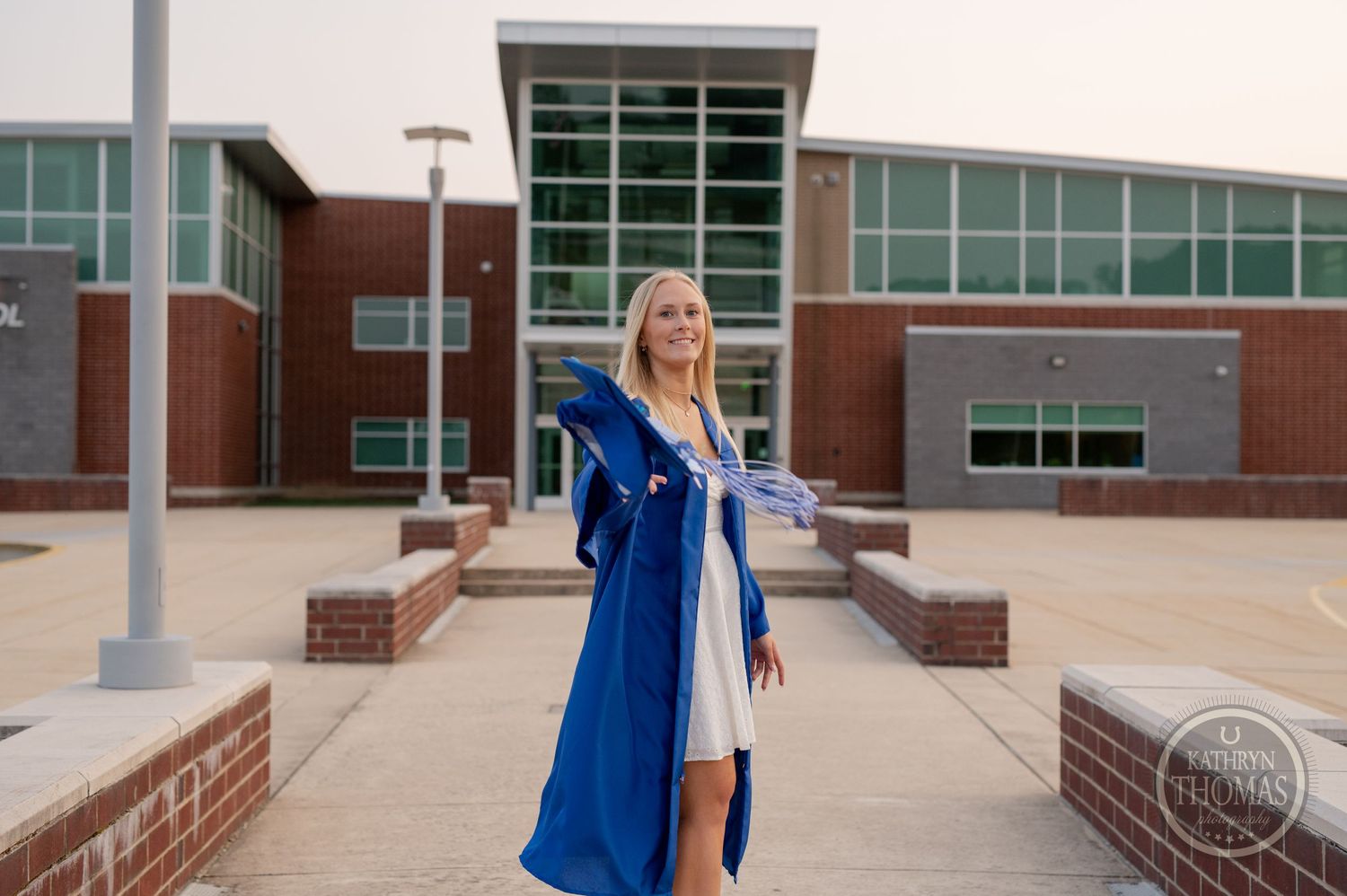 Graduate in blue commencement regalia poses confidently outside modern brick school building.
