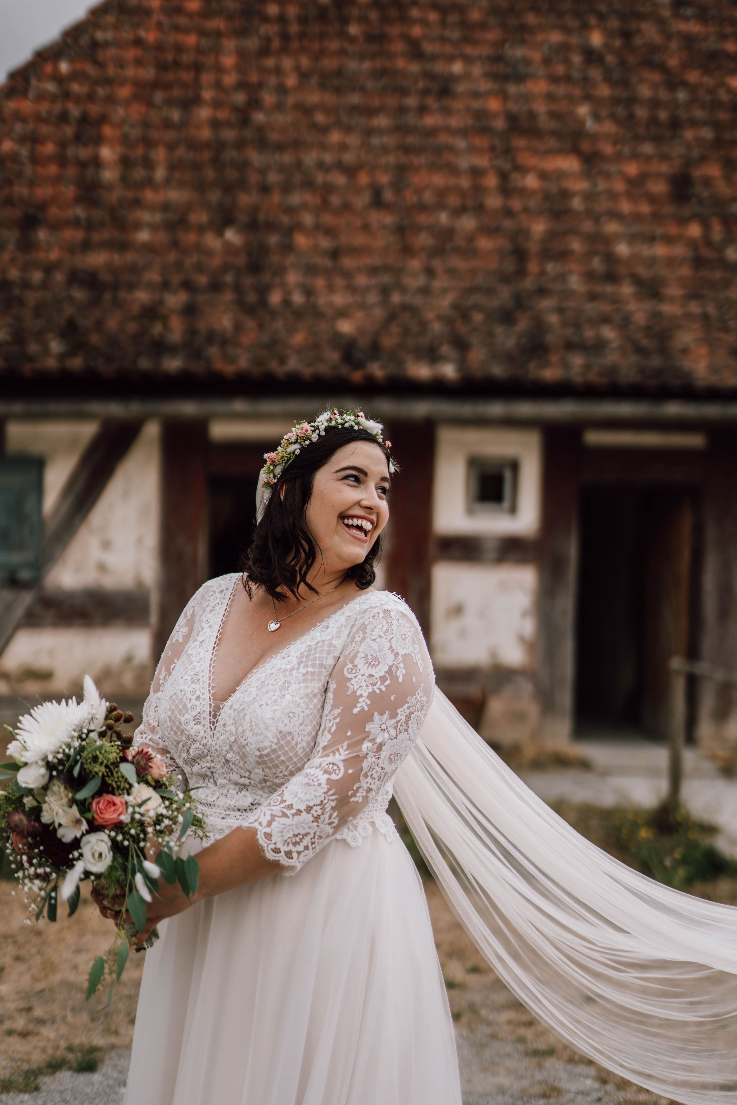Beautiful bride in lace dress with flowing veil poses against rustic backdrop.