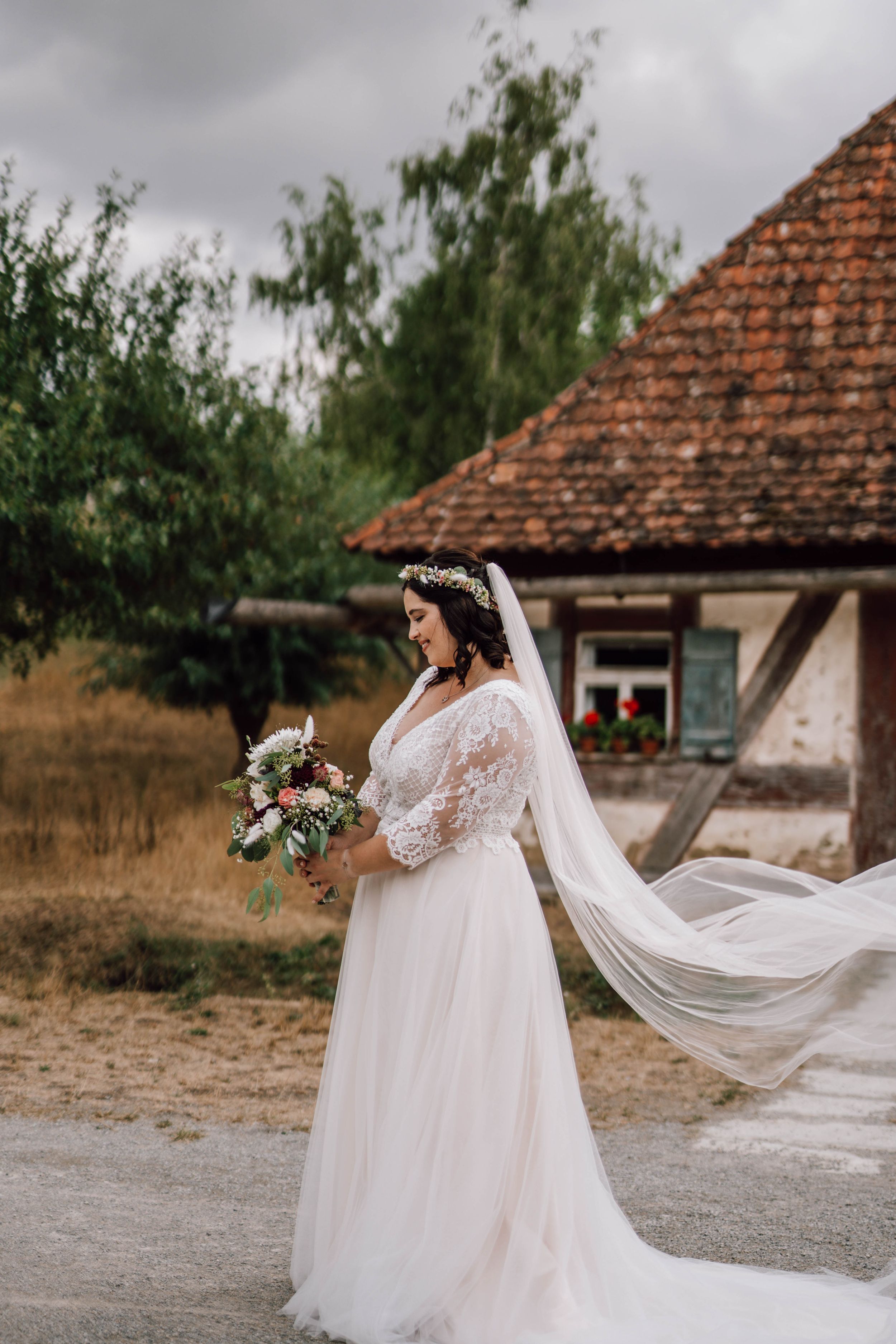Bride's flowing white dress and veil catch the wind in a rustic outdoor setting.