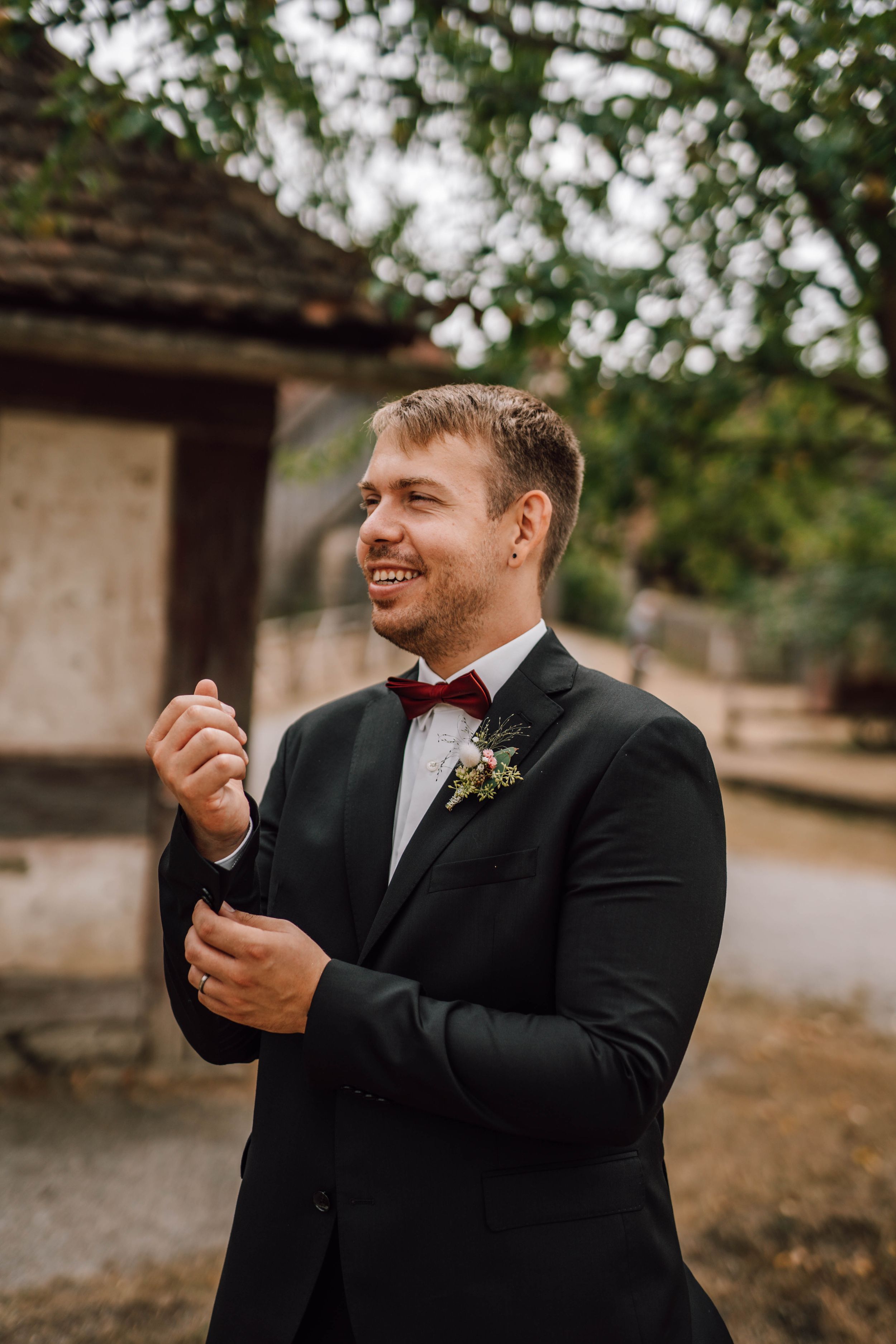 Groom in black tuxedo and burgundy bow tie laughs during outdoor ceremony.