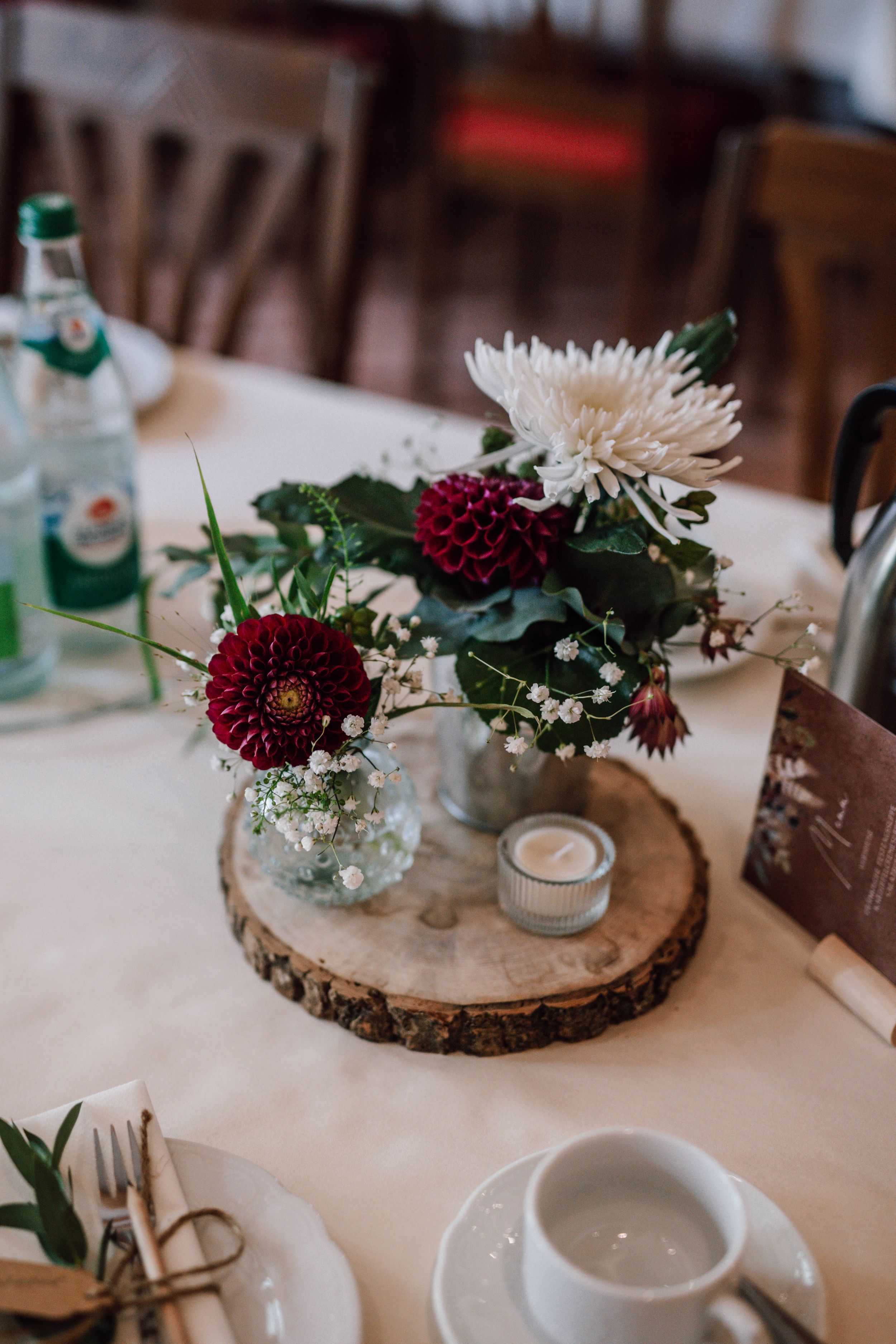 Rustic wedding centerpiece with burgundy and white flowers on wood slice with glass vase and table settings.