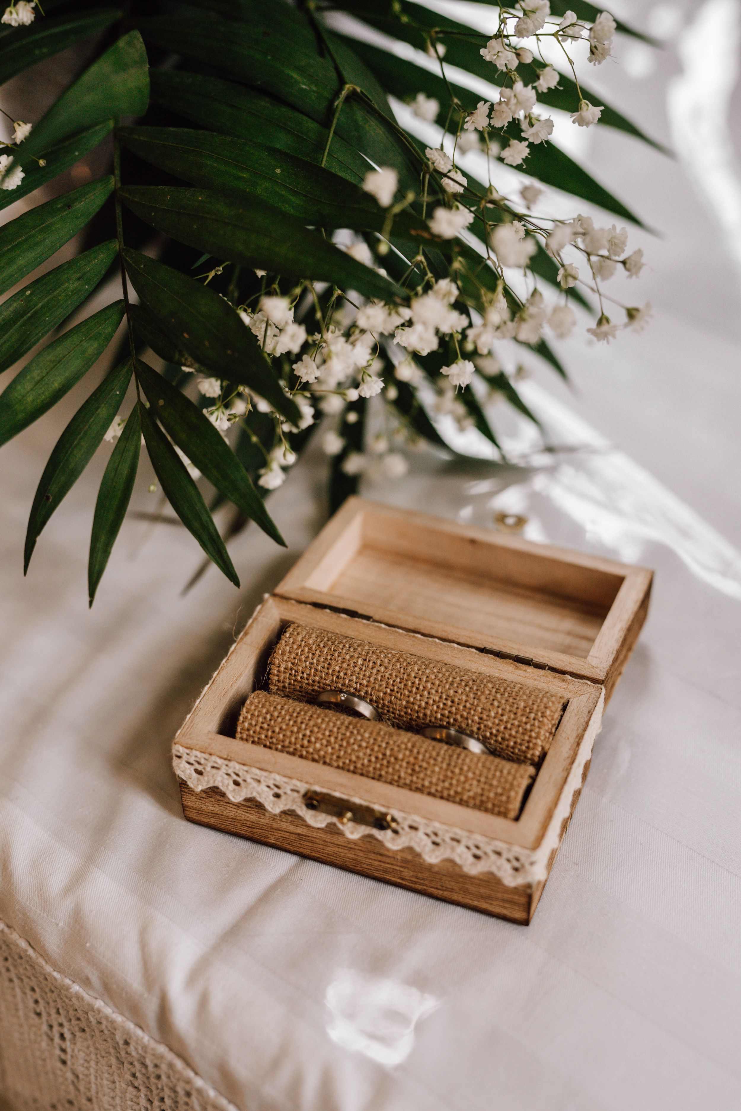 Wedding rings displayed in a rustic wooden box with burlap and lace under tropical leaves.