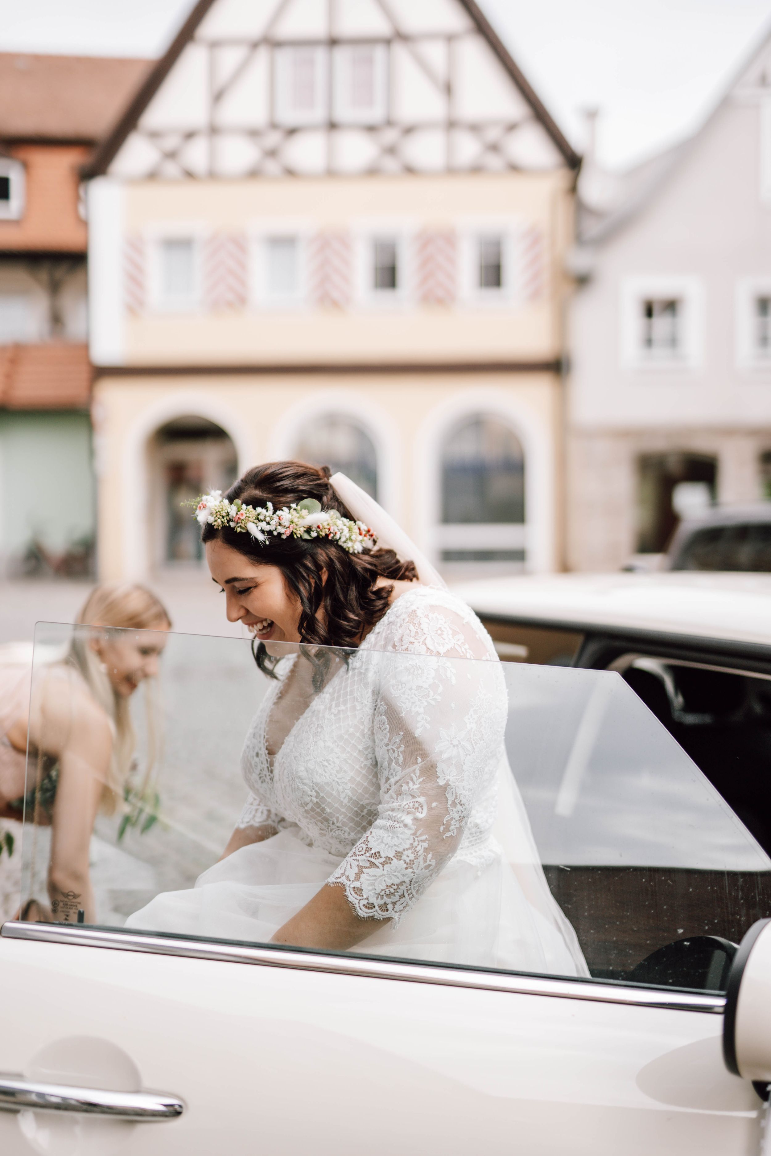 A bride in a lace gown enters a classic white wedding car.