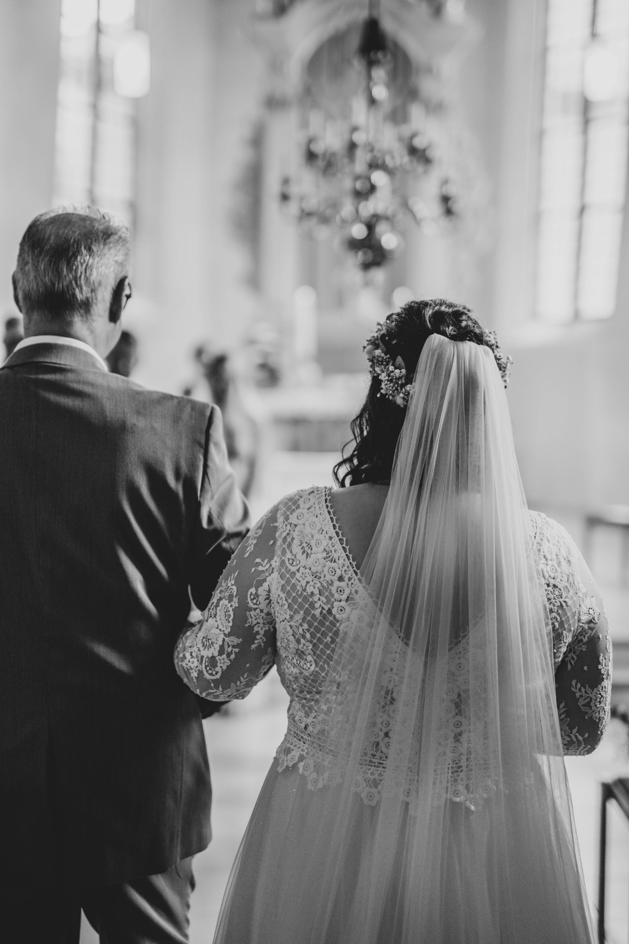 A bride in a lace dress and flowing veil stands in a sunlit church interior.