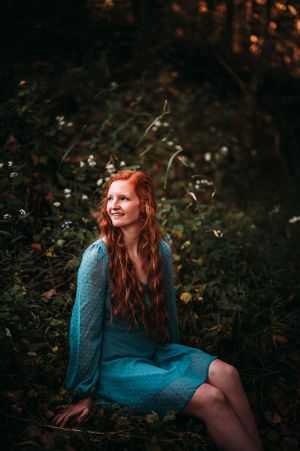 Smiling young woman with long red hair sitting among wildflowers wearing a teal dress in forest.