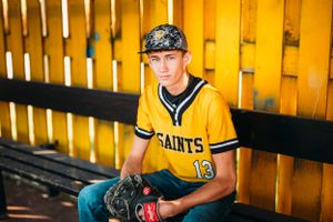 Teen baseball player in yellow Saints jersey number 13 sitting in dugout with glove.