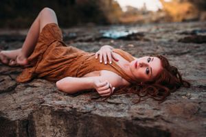 Red-haired woman in brown dress lying on rocky surface near water at dusk.