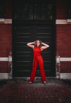 Smiling woman in a red jumpsuit poses confidently in front of a black door on a brick building.