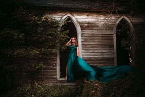 Woman in flowing teal gown poses dramatically in the arched window of an abandoned wooden building.
