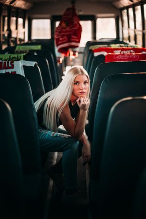 Blonde woman sits pensively on a school bus seat, gazing forward among rows of empty green seats.