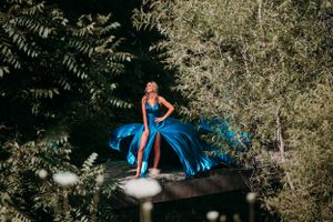 Blonde woman in a stunning blue satin gown stands atop a structure surrounded by lush green trees.