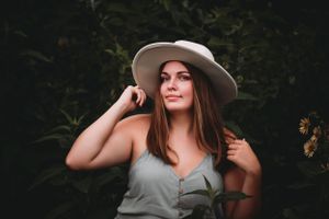 Young woman in a wide-brim hat and sage green top poses among lush green foliage in a senior portrait.