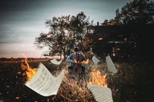 Burning sheet music flies through flames in a dramatic outdoor scene with a cellist performing near a tree at dusk.