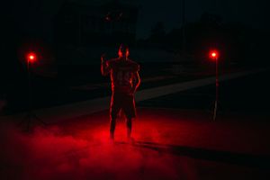 Muscular man poses in red smoke and light at night, flanked by two glowing red torches.