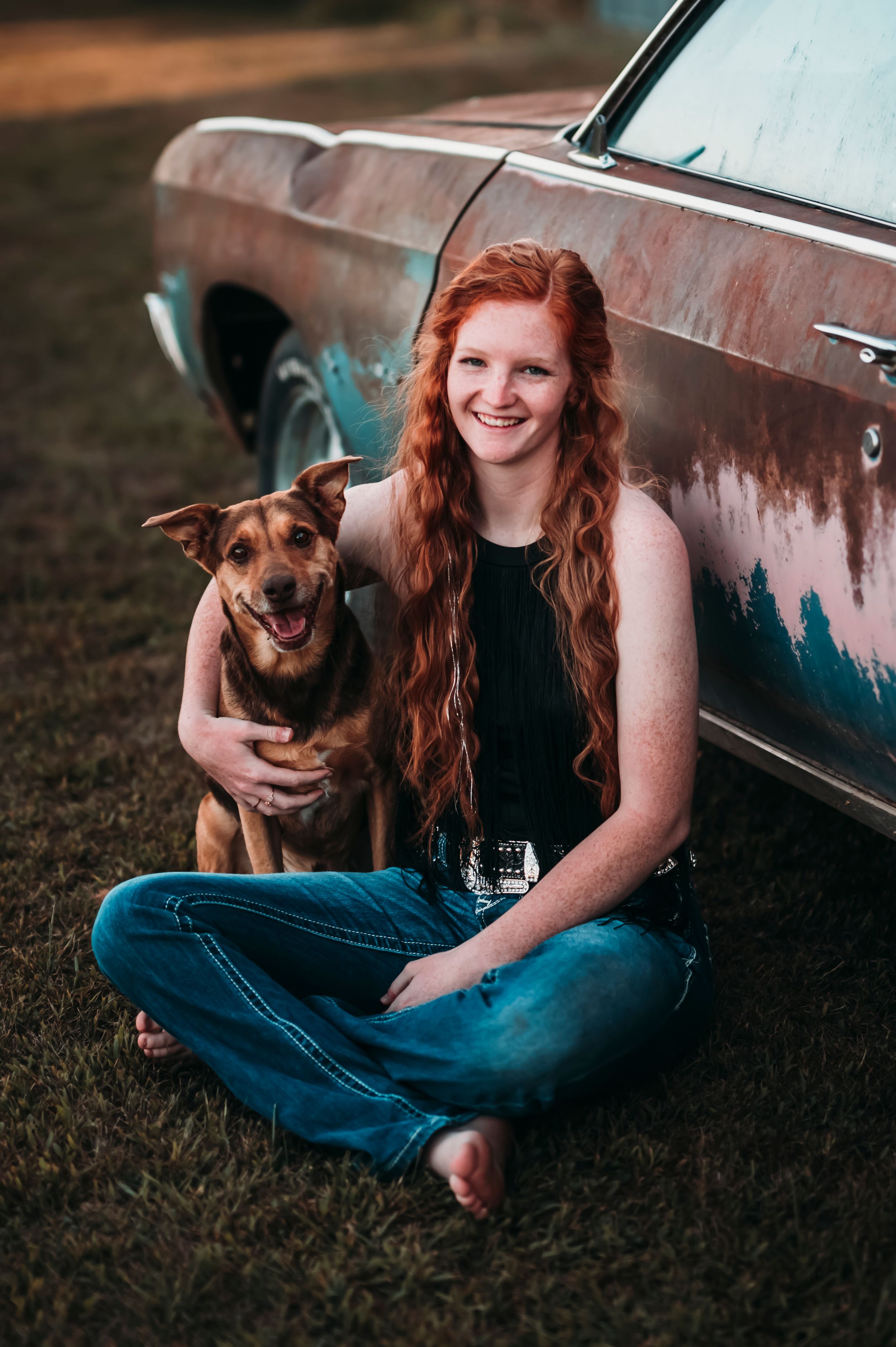 Smiling redhead woman sitting with brown dog beside rustic vintage car outdoors.