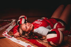 Young female volleyball player in red uniform lying on court surrounded by volleyballs smiling.